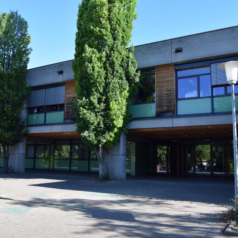 Ein modernes Schulgebäude mit großen Fenstern und Bäumen im Vordergrund.